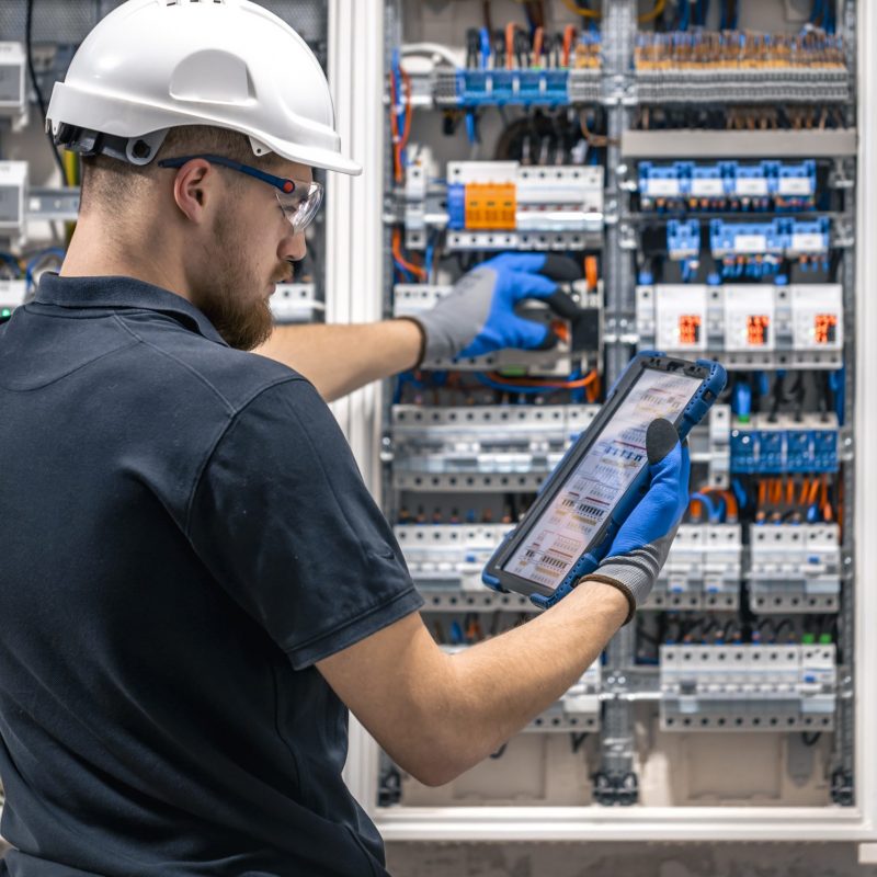 Electrical technician working in a switchboard with fuses, uses a tablet. Electrical technician looking focused while working in a switchboard with fuses.